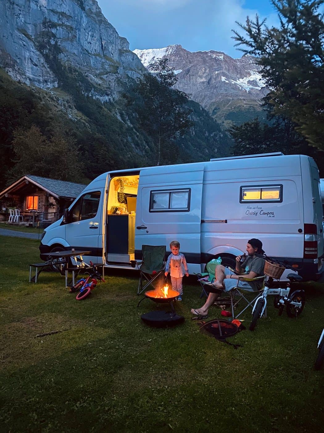 A Camper Camper van at a mountain campsite at dusk, with the family round a fire pit